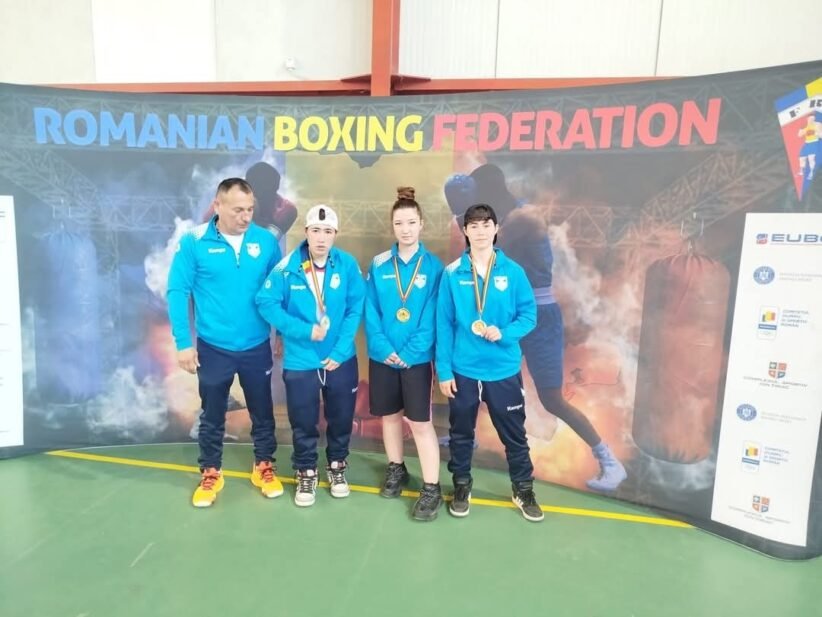 Coach and three young boxers in blue jackets pose with medals in front of a Romanian Boxing Federation banner indoors.