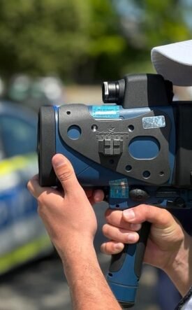 Police officer using a handheld speed radar gun, with a police car blurred in the background.