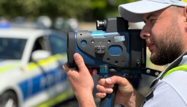 Police officer using a handheld speed radar gun, with a police car blurred in the background.