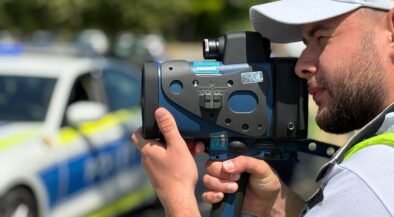 Police officer using a handheld speed radar gun, with a police car blurred in the background.