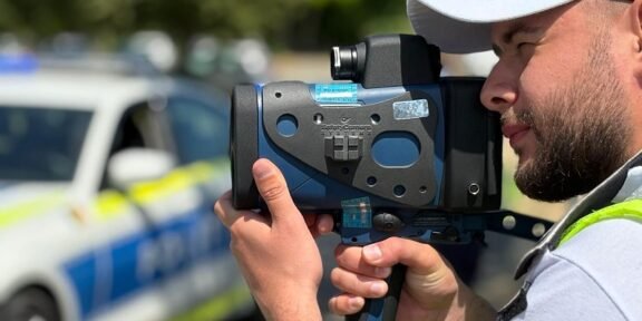 Police officer using a handheld speed radar gun, with a police car blurred in the background.