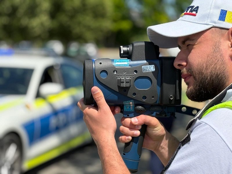 Police officer using a handheld speed radar gun, with a police car blurred in the background.