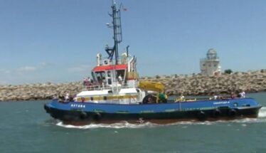 Blue tugboat Astana with a red-roofed cabin and yellow crane, sailing near a rocky breakwater on a sunny day in calm water.