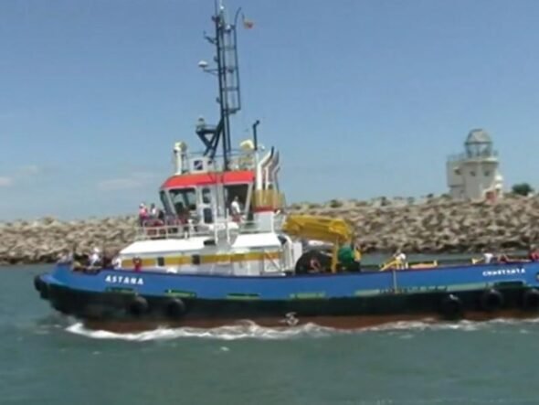 Blue tugboat Astana with a red-roofed cabin and yellow crane, sailing near a rocky breakwater on a sunny day in calm water.