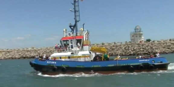 Blue tugboat named Astana speeds through a harbor with a rocky breakwater and a white lighthouse-like structure in the distance, several people on deck visible.