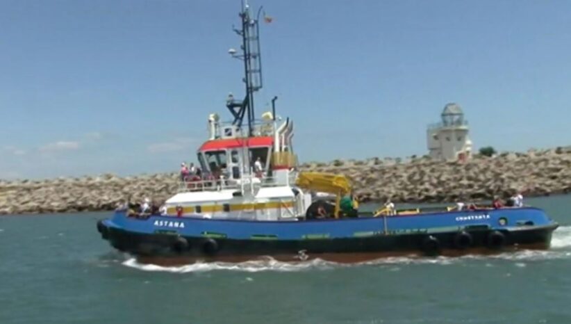 Blue tugboat named Astana speeds through a harbor with a rocky breakwater and a white lighthouse-like structure in the distance, several people on deck visible.