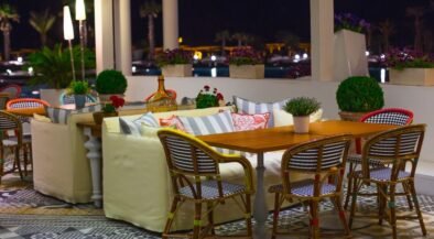 Nighttime outdoor cafe with wooden table and wicker chairs, cream couches, and potted plants on patterned tiled floor.
