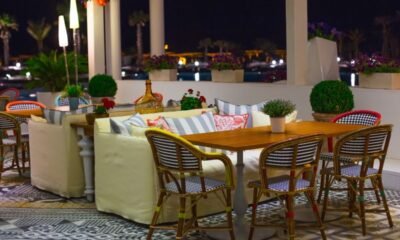 Nighttime outdoor cafe with wooden table and wicker chairs, cream couches, and potted plants on patterned tiled floor.