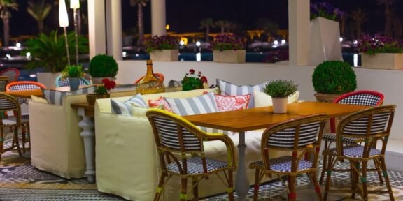 Nighttime outdoor cafe with wooden table and wicker chairs, cream couches, and potted plants on patterned tiled floor.