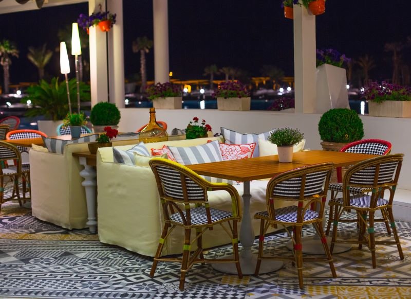 Nighttime outdoor cafe with wooden table and wicker chairs, cream couches, and potted plants on patterned tiled floor.