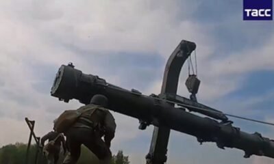 Two soldiers in camouflage stand beside a large rocket or missile being lifted by a crane outdoors, preparing it for installation or maintenance.