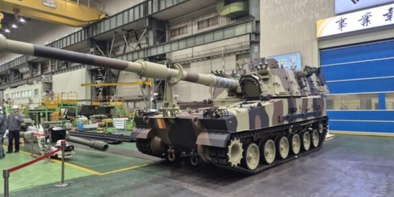 Camouflage military tank on rails in an indoor maintenance facility, long barrel extending forward.