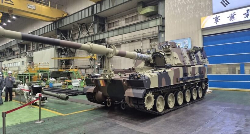 Camouflage military tank on rails in an indoor maintenance facility, long barrel extending forward.