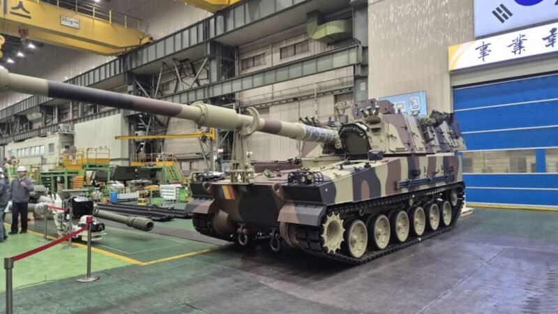 Camouflage military tank on rails in an indoor maintenance facility, long barrel extending forward.