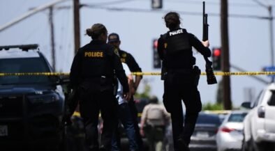 Police officers at a crime scene behind yellow tape, one officer holds a long firearm while another stands near a parked SUV.