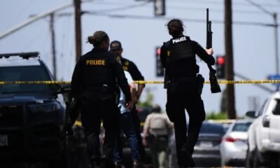 Police officers at a crime scene behind yellow tape, one officer holds a long firearm while another stands near a parked SUV.