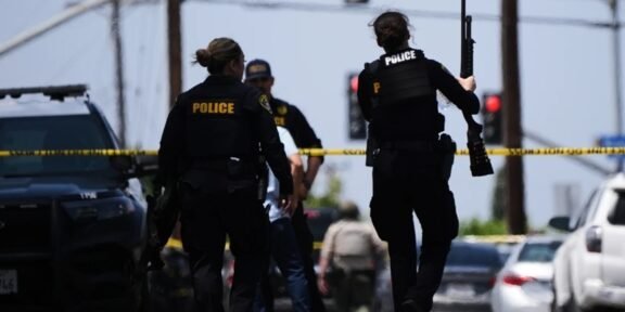 Police officers at a crime scene behind yellow tape, one officer holds a long firearm while another stands near a parked SUV.