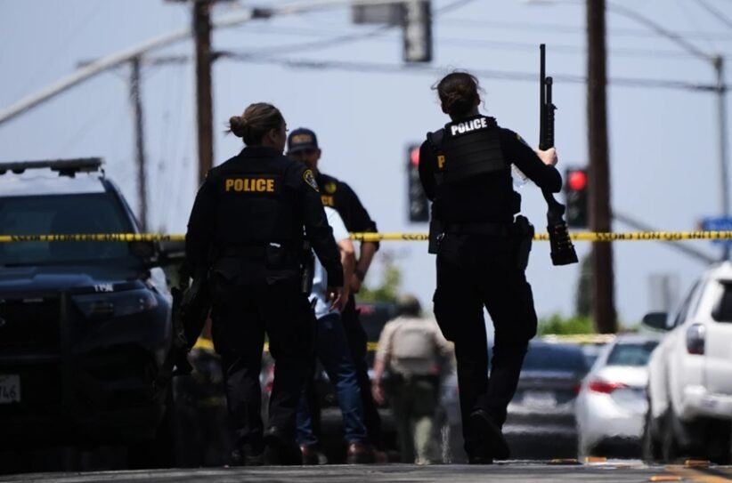 Police officers at a crime scene behind yellow tape, one officer holds a long firearm while another stands near a parked SUV.