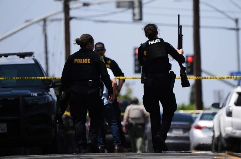 Police officers at a crime scene behind yellow tape, one officer holds a long firearm while another stands near a parked SUV.