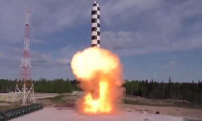 Rocket launching vertically from a pad with a bright orange flame plume and smoke, beside a tall support tower.