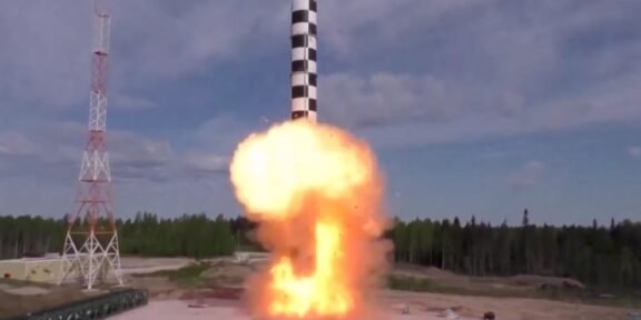 Rocket launching vertically from a pad with a bright orange flame plume and smoke, beside a tall support tower.