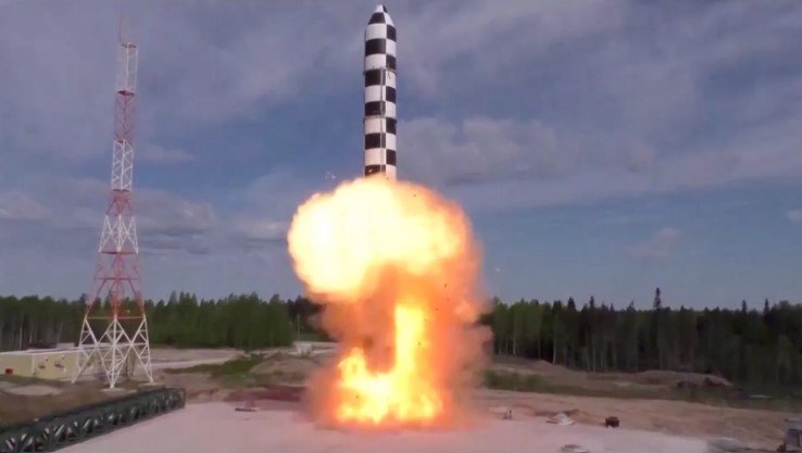 Rocket launching vertically from a pad with a bright orange flame plume and smoke, beside a tall support tower.