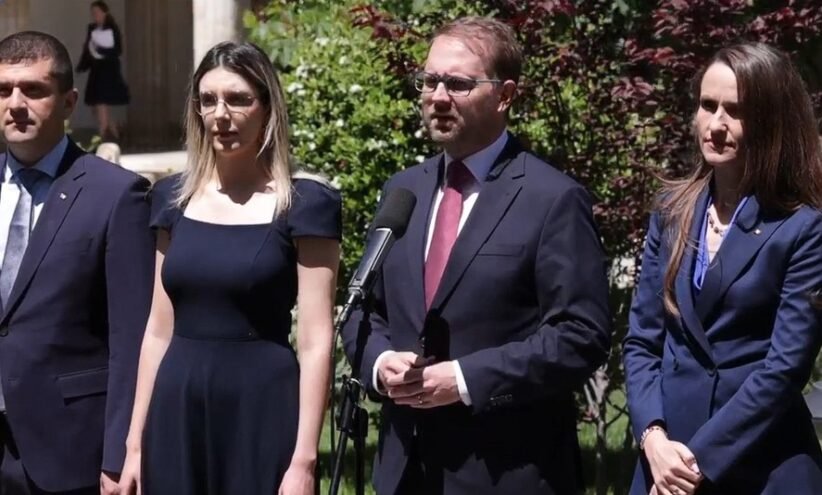 Four professionals in business attire stand outdoors; a man speaks into a microphone while three others listen nearby.