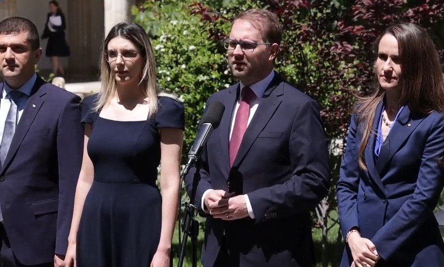 Four professionals in business attire stand outdoors; a man speaks into a microphone while three others listen nearby.