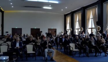 Audience seated in a large conference hall, awaiting a presentation; natural light from tall windows on the right side.