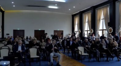 Audience seated in a large conference hall, awaiting a presentation; natural light from tall windows on the right side.