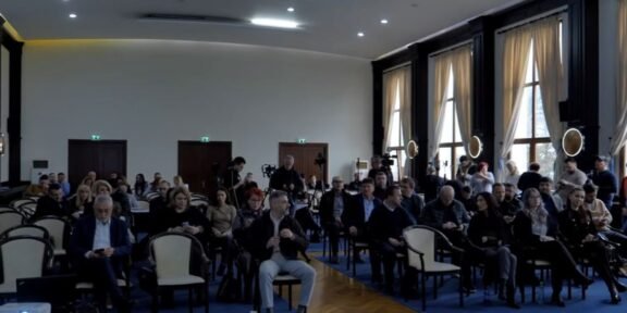 Audience seated in a large conference hall, awaiting a presentation; natural light from tall windows on the right side.