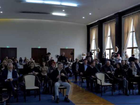 Audience seated in a large conference hall, awaiting a presentation; natural light from tall windows on the right side.