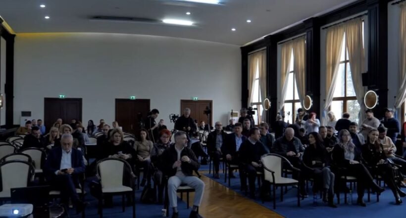 Audience seated in a large conference hall, awaiting a presentation; natural light from tall windows on the right side.