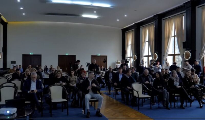 Audience seated in a large conference hall, awaiting a presentation; natural light from tall windows on the right side.