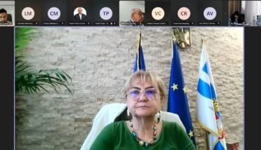 Framed woman in a green blouse wearing glasses, seated in a conference setting with European and flag banners behind her.
