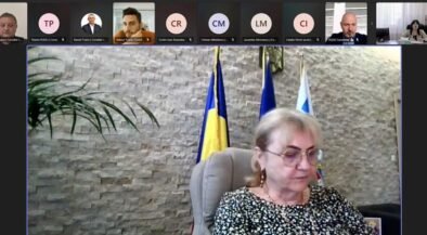 Video conference screenshot: a woman in a patterned blouse sits at a desk, with flags behind her and participants along the top thumbnails.
