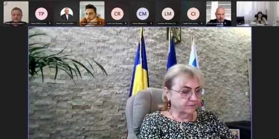 Video conference screenshot: a woman in a patterned blouse sits at a desk, with flags behind her and participants along the top thumbnails.