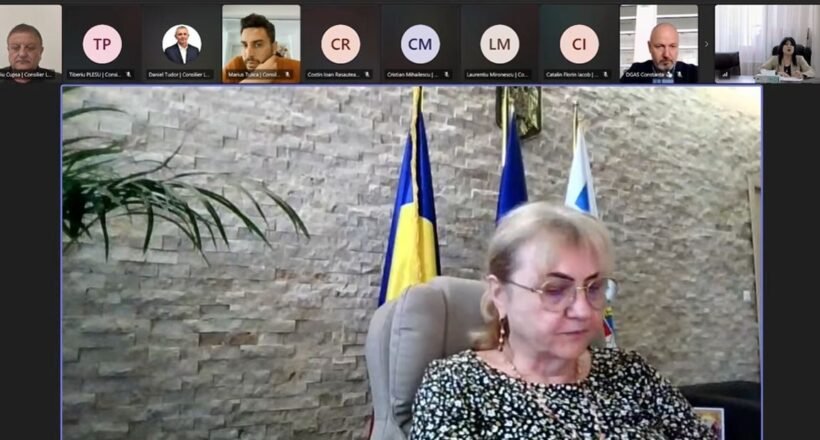 Video conference screenshot: a woman in a patterned blouse sits at a desk, with flags behind her and participants along the top thumbnails.