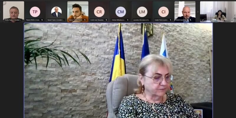 Video conference screenshot: a woman in a patterned blouse sits at a desk, with flags behind her and participants along the top thumbnails.