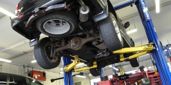 Car raised on a hydraulic lift in an auto repair shop, viewed from underneath, showing the exhaust, rear axle, suspension, and tires with blue and yellow lift arms overhead.