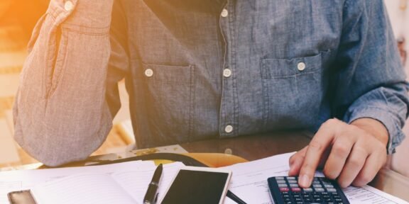 Person in a denim shirt calculating finances at a desk with a calculator, smartphone, and notebook open in front of them