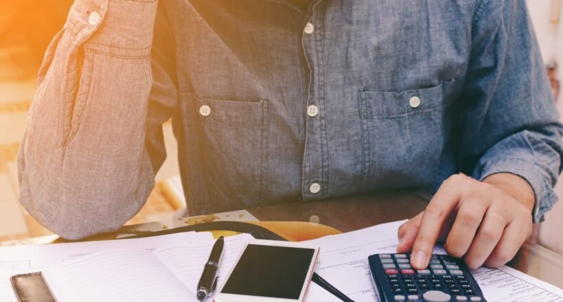 Person in a denim shirt calculating finances at a desk with a calculator, smartphone, and notebook open in front of them