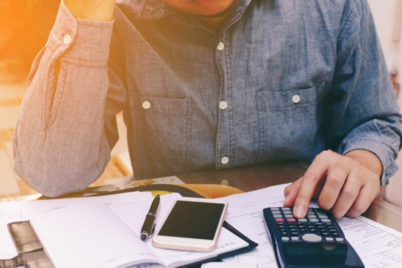 Person in a denim shirt calculating finances at a desk with a calculator, smartphone, and notebook open in front of them