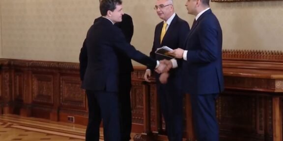 Four men in suits shake hands in a formal, wood-paneled room while two others stand nearby, one holding a booklet.