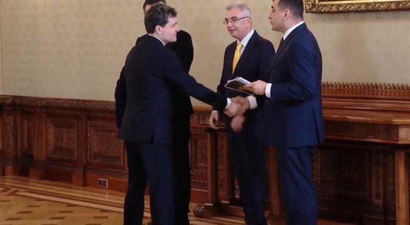 Four men in suits shake hands in a formal, wood-paneled room while two others stand nearby, one holding a booklet.