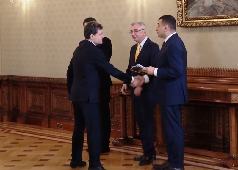 Four men in suits shake hands in a formal, wood-paneled room while two others stand nearby, one holding a booklet.