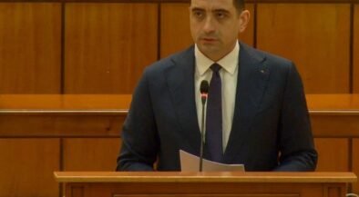 Man in a dark suit delivering remarks at a wooden podium with a microphone in a parliamentary chamber. The podium reads 'Camera Deputaților'.