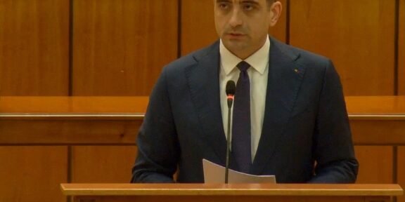 Man in a dark suit delivering remarks at a wooden podium with a microphone in a parliamentary chamber. The podium reads 'Camera Deputaților'.
