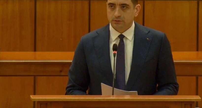 Man in a dark suit delivering remarks at a wooden podium with a microphone in a parliamentary chamber. The podium reads 'Camera Deputaților'.