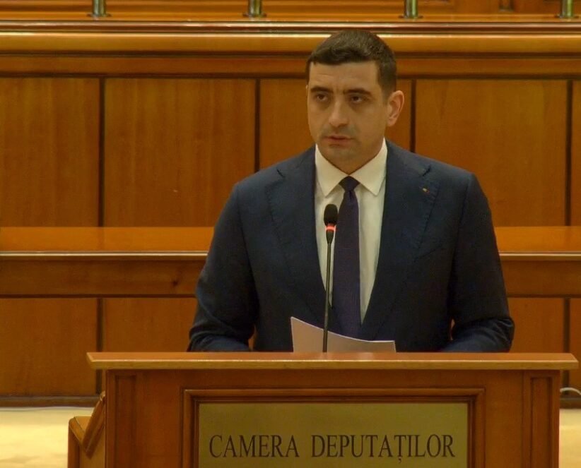 Man in a dark suit delivering remarks at a wooden podium with a microphone in a parliamentary chamber. The podium reads 'Camera Deputaților'.